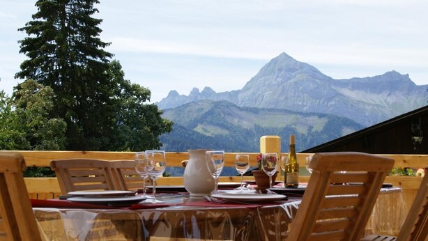 Ein Esstisch mit Stühlen, einer Vase, Weingläsern und einer Flasche, mit Blick auf eine Berglandschaft.