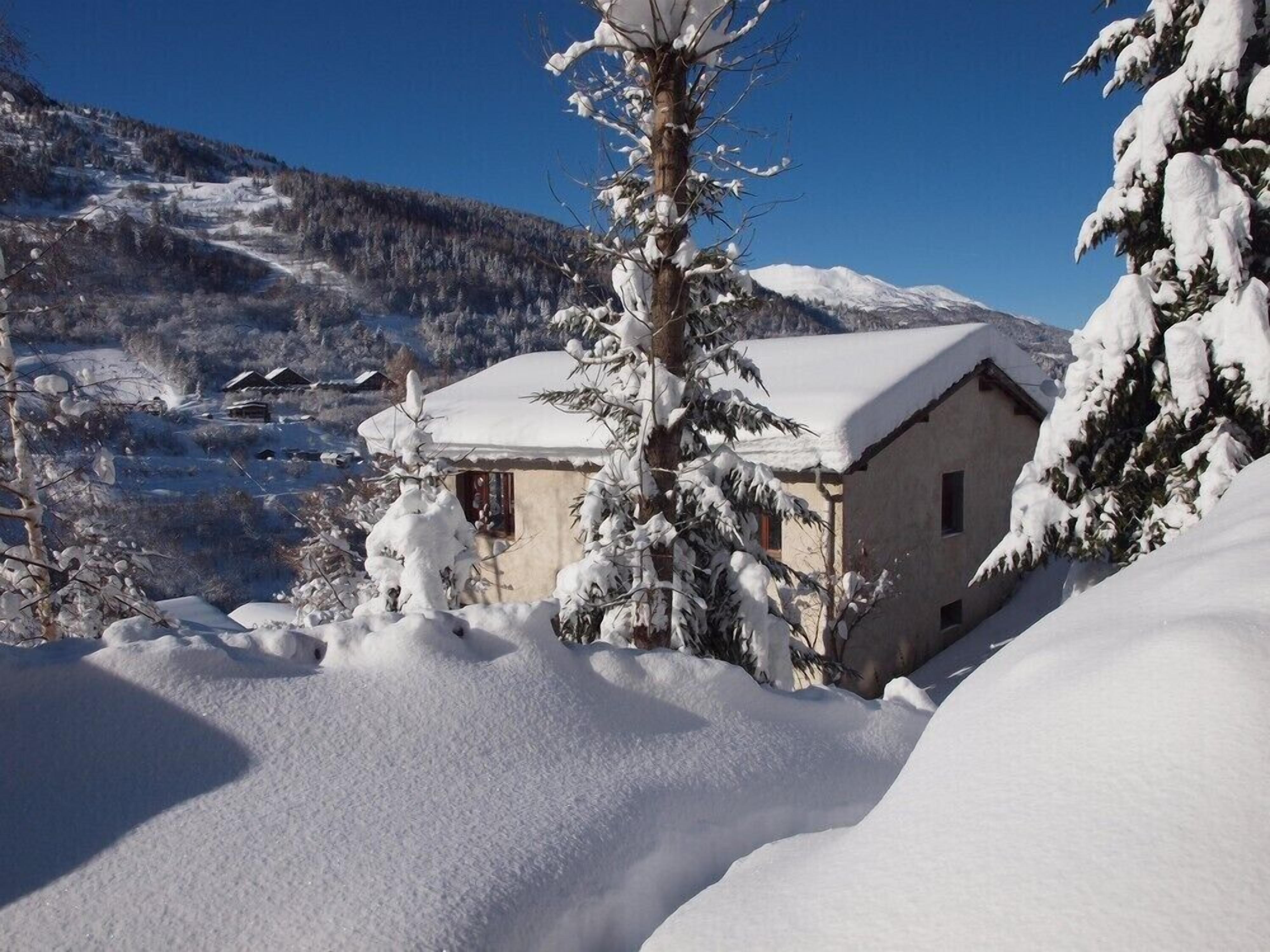 Paisagem montanhosa coberta de neve com um pequeno edifício rodeado por árvores.