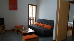 A small, cozy room with a sofa, a wooden table, a television, and a door leading to another room.