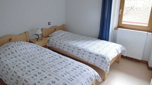 A small, clean room with two wooden beds, a nightstand, a window with blue curtains, and a radiator.