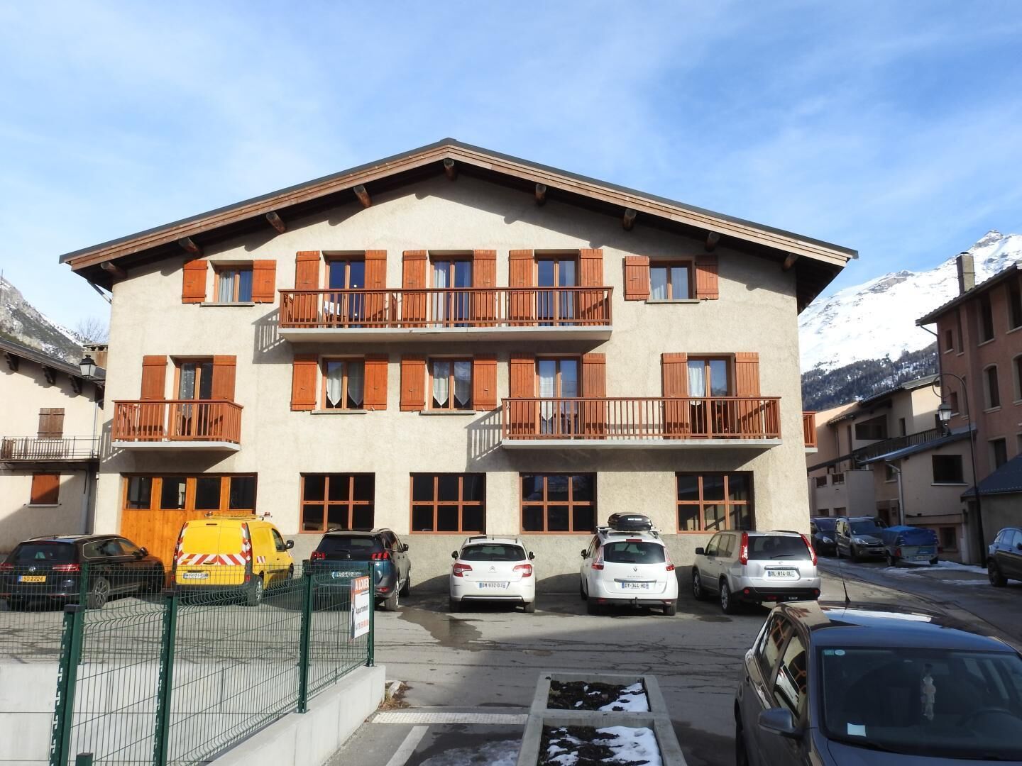 Un edificio de varios pisos con balcones, un estacionamiento con coches y montañas al fondo.