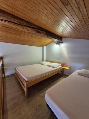 Uma cama de solteiro com cabeceira de madeira, mesa de cabeceira e luminária fixada na parede em um quarto com vigas de madeira no teto.