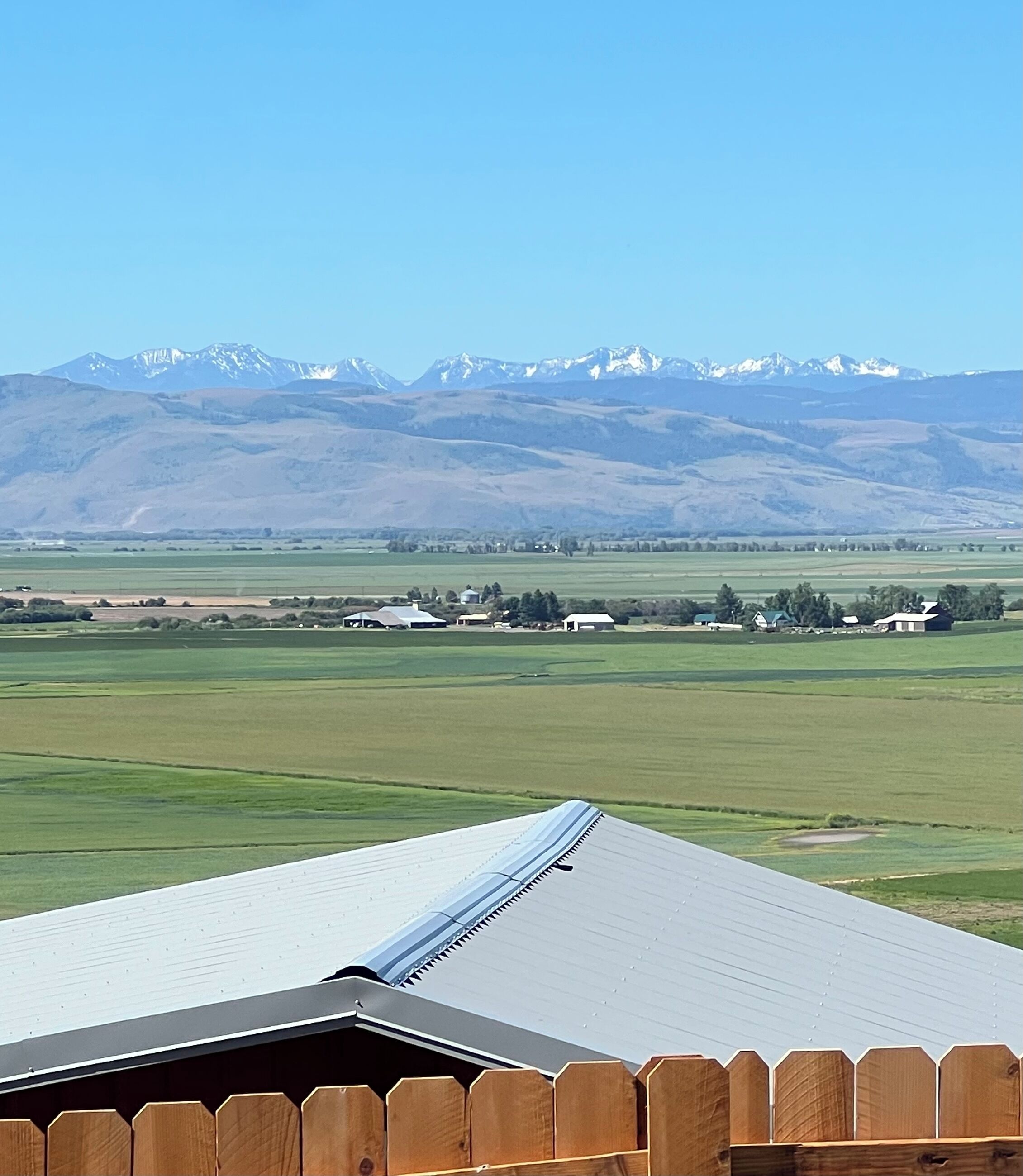 Une vue d’un paysage rural, avec une clôture en bois au premier plan, un champ verdoyant et une chaîne de montagnes en arrière-plan.