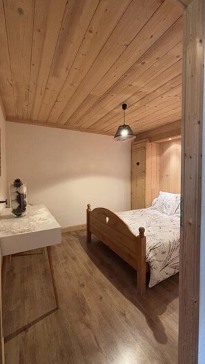 Une chambre avec un plafond en bois, un lit recouvert d’une couvre-lit à motifs floraux, une table de chevet avec un vase et une armoire en bois.