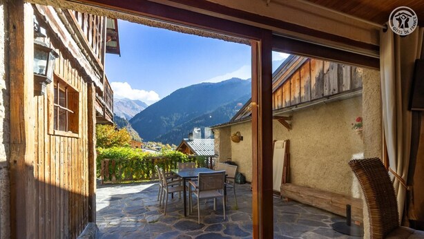 A stone-floored patio with a table and chairs, a wicker chair, and a view of mountains.