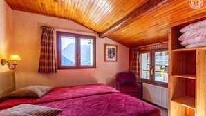 A cozy room with wooden ceiling and walls, a bed with red bedding, a chair, and a shelf with pillows.