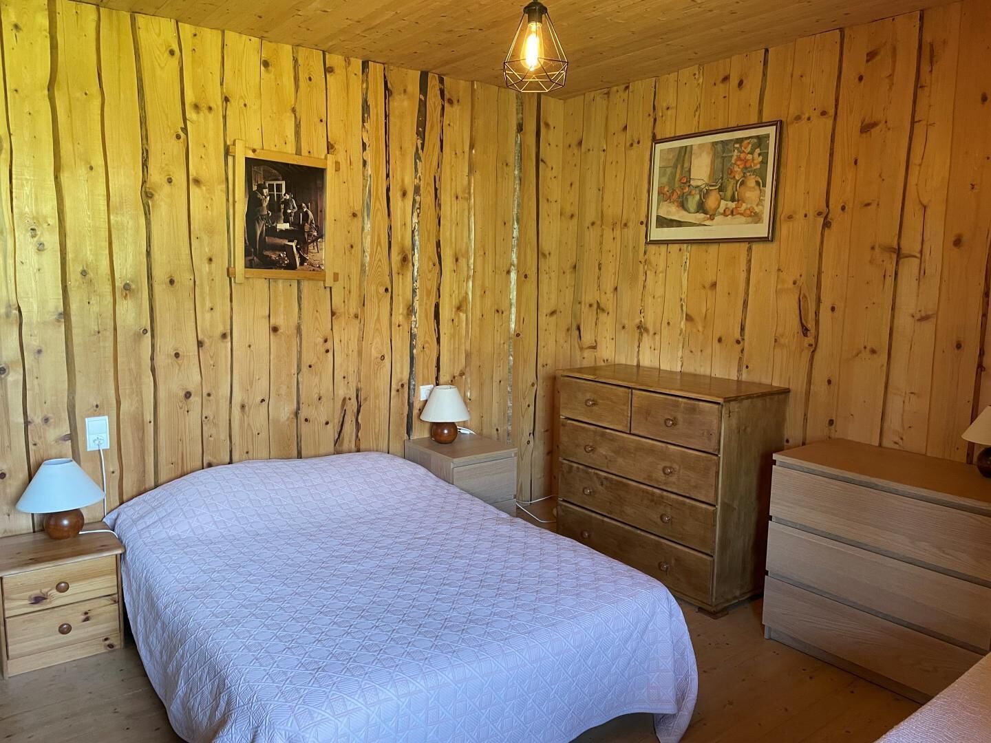 Une pièce aux murs lambrissés, comprenant un lit, deux tables de chevet, une commode et un luminaire suspendu.