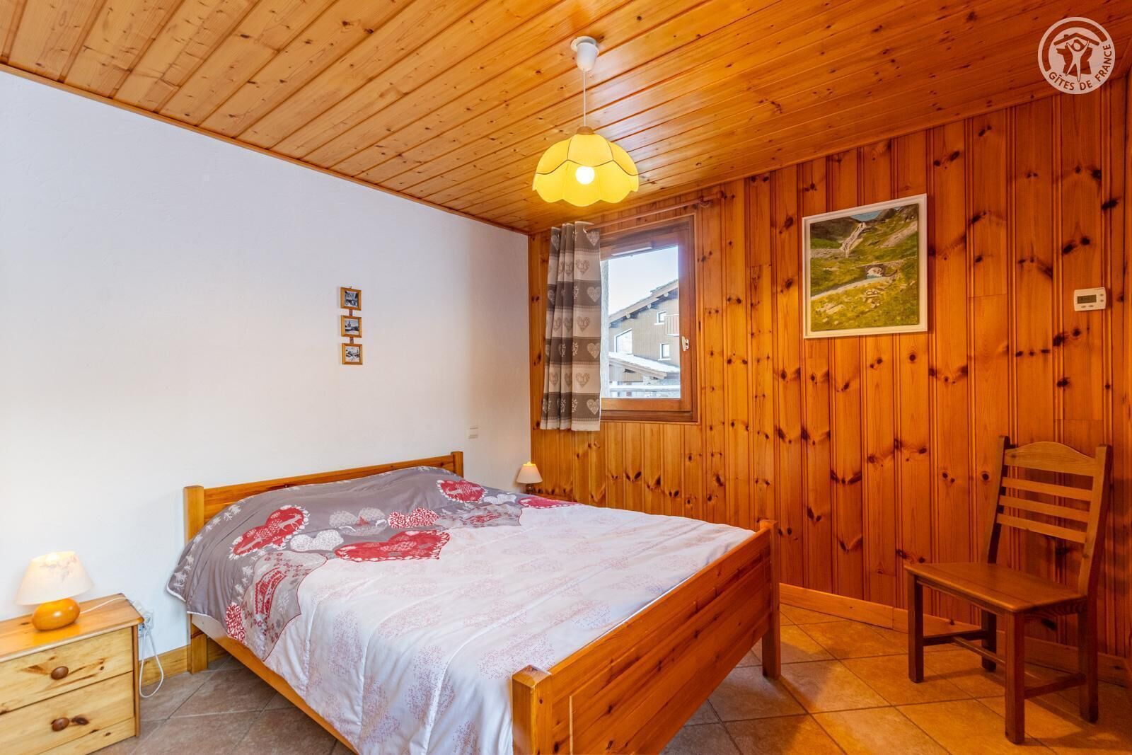 Une pièce aux murs lambrés, comprenant un lit, une chaise, une table de chevet, une fenêtre avec des rideaux et un thermostat fixé au mur.