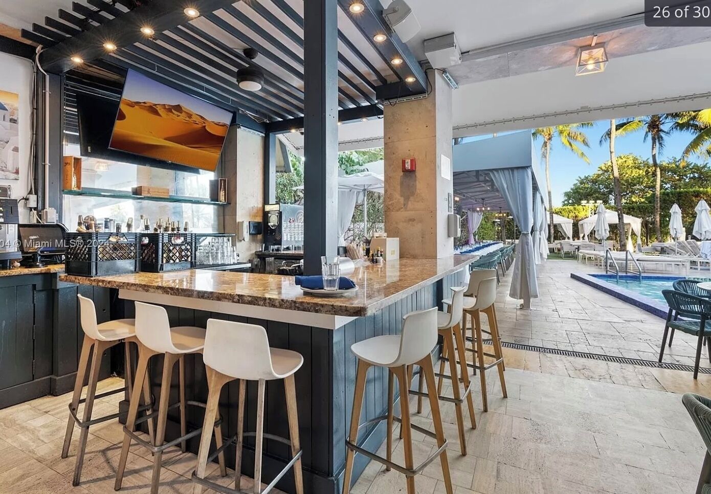 A bar area with a poolside view, featuring a bar counter, stools, and a large screen.