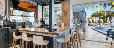 A bar area with a poolside view, featuring a bar counter, stools, and a large screen.