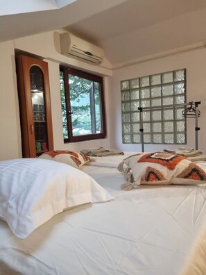 A hotel room with two beds, an air conditioning unit, a window with a view of greenery, and a door leading to a bathroom.