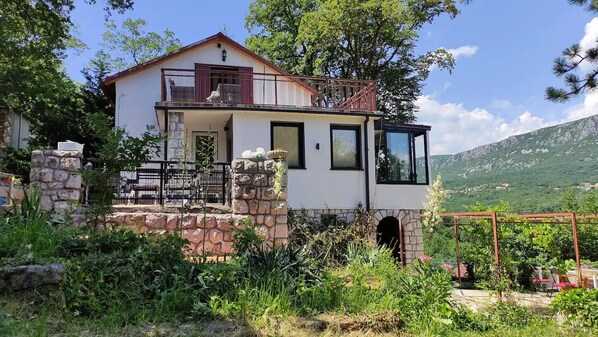 A two-story house with a stone foundation, a balcony, and a view of a mountainous landscape.