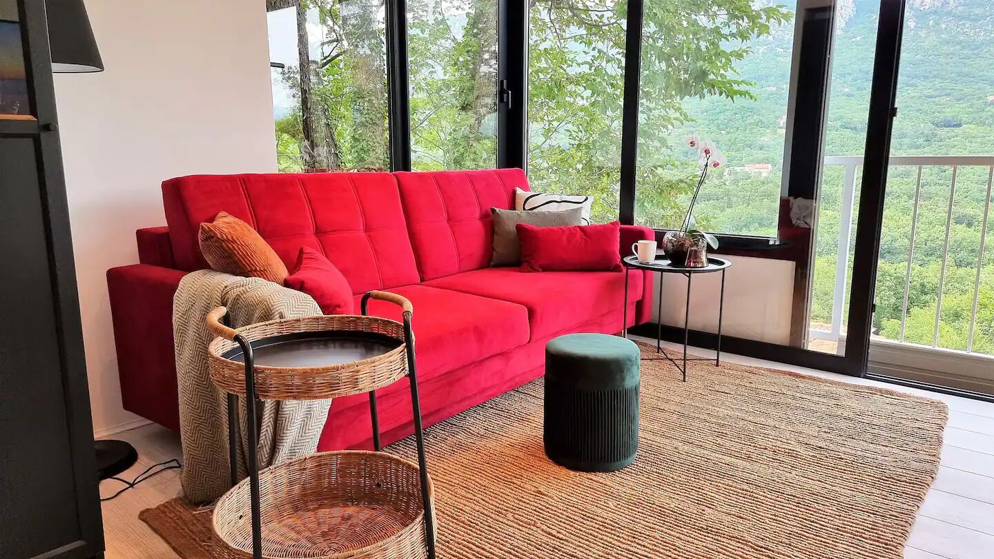 Un divano rosso con cuscini, un tavolino in vimini, un poggiapiedi e vista sul verde attraverso ampie finestre.