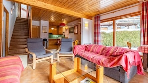 Een houten interieur met een bank, stoelen, een glazen salontafel, een trap en een keukenhoek.