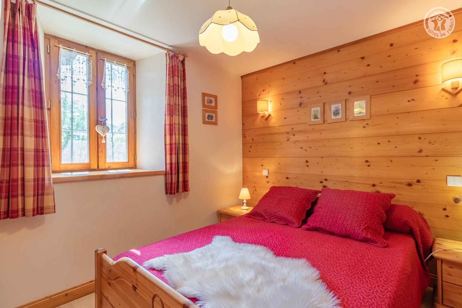 A bedroom with a wooden headboard, a bed with red bedding, a white fluffy rug, a window with curtains, and wall-mounted lights.