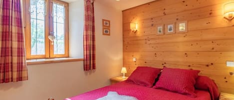 A bedroom with a wooden headboard, a bed with red bedding, a white fluffy rug, a window with curtains, and wall-mounted lights.