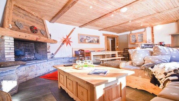 Ein rustikales Interieur mit Kamin aus Stein, Holzbalkendecke, Essbereich mit Tisch und Stühlen sowie eine gemütliche Sitzecke mit einer Couch.