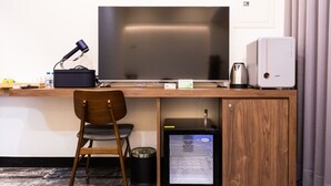 Ein kompakter Arbeitsbereich im Hotelzimmer mit Schreibtisch, Stuhl, Fernseher, Kaffeemaschine und einem kleinen Kühlschrank.