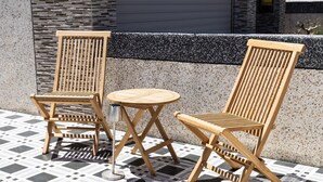 Zwei Holzstühle und ein kleiner runder Tisch auf einer gefliesten Terrasse mit einer Steinmauer im Hintergrund.
