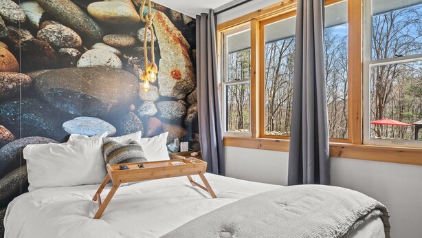 Ein Schlafzimmer mit einem großen Bett, einem Nachttisch aus Holz, einer Wandmalerei von Steinen und einem Fenster mit Blick auf Bäume.