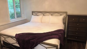 Un lit avec un matelas blanc et une couverture violette, une fenêtre donnant sur des arbres, et une commode en bois foncé avec des tiroirs.
