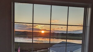 Ein Zimmer mit großem Fenster mit Blick auf einen Sonnenuntergang über einem Gewässer und ein Haus.