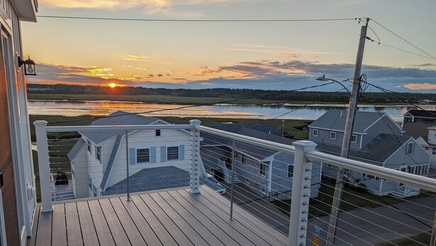 Eine Holzterrasse mit Geländer, die auf ein Wohngebiet und ein Gewässer im Sonnenuntergang blickt.