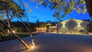 Um edifício de pedra com entrada por um caminho de cascalho, rodeado por árvores e iluminação exterior.