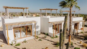 Un hotel moderno y minimalista con paredes blancas y pérgolas de madera. Cada unidad tiene una puerta de vidrio que conduce a una habitación privada. La propiedad está rodeada por un paisaje desértico con cactus y palmeras.