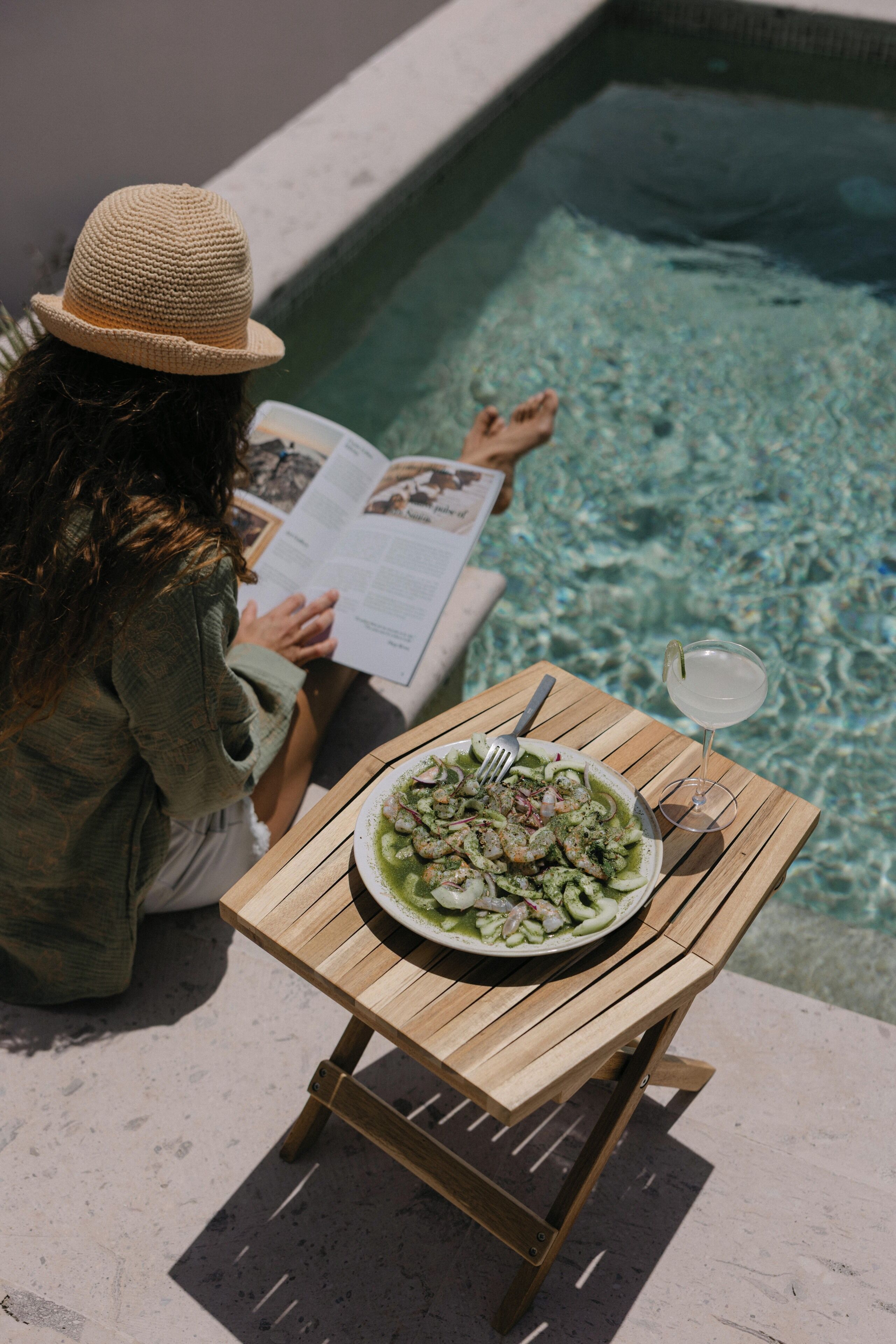 Eine Person liest an einem Tisch am Pool, neben einem Teller Essen und einem Getränk.