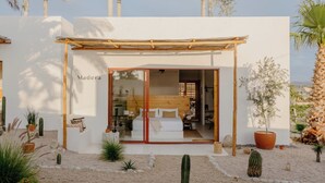 Un edificio moderno con entrada tipo pérgola de madera, un dormitorio visible con cama y sofá, y un jardín desértico con cactus y plantas.