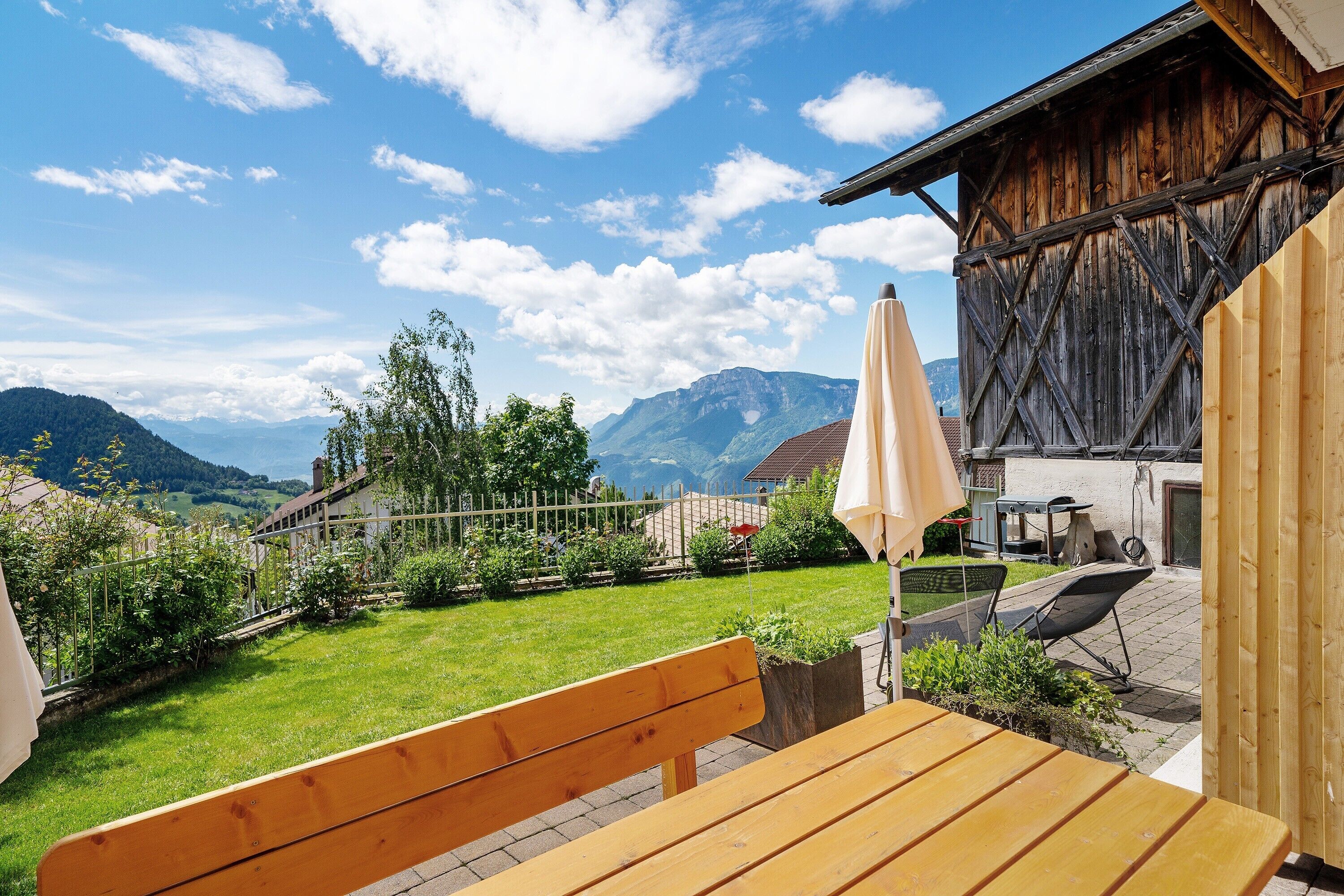 A wooden bench and table in a garden with a mountain view, a wooden building, and a white umbrella.