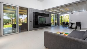 A modern living room with a flat-screen TV, a gray sofa, and a dining area with chairs. - Villa 'Green Garden' with Private Terrace, Wi-Fi and Air Conditioning (Carlentini)