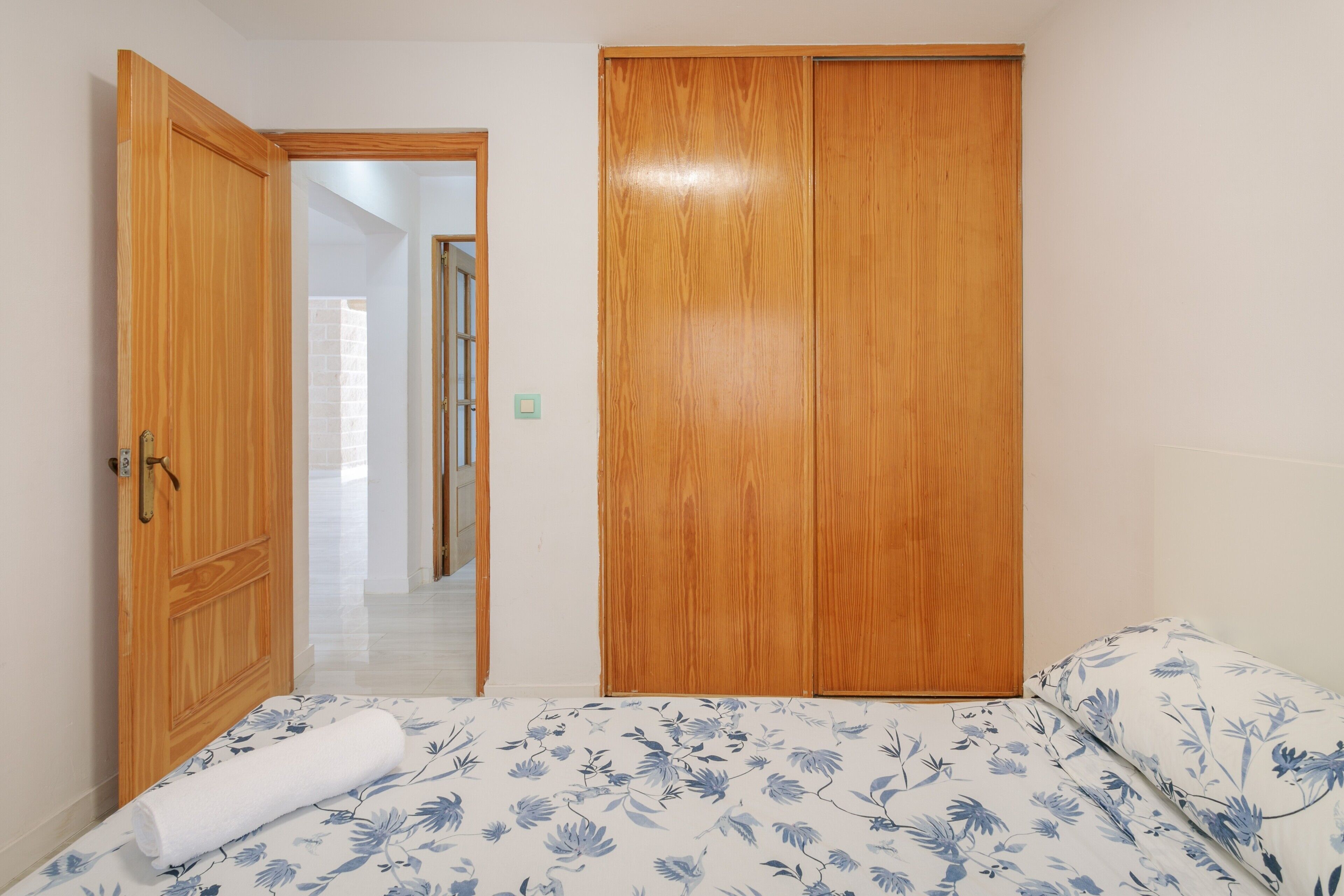 A bedroom with a wooden wardrobe, a bed with floral bedding, and a hallway leading to another room.