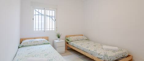 A room with two wooden beds, a white nightstand, a window with bars, and a small potted plant.