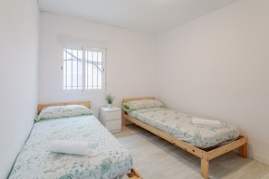 Ein Zimmer mit zwei hölzernen Betten, einem weißen Nachttisch, einem Fenster mit Gitterstäben und einer kleinen Topfpflanze.