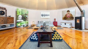 A room with a wooden floor, a central table, a sofa, a bed, a fireplace, and various wall decorations including a Buddha painting and a stone arrangement.