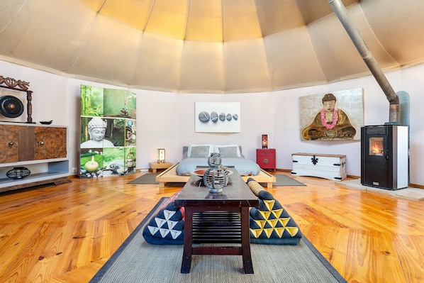 A room with a wooden floor, a central table, a sofa, a bed, a fireplace, and various wall decorations including a Buddha painting and a stone arrangement.
