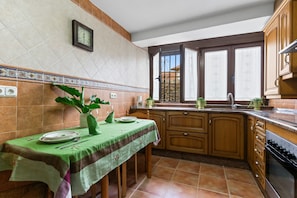 A kitchen with a dining table set, wooden cabinets, a window with a view, and a clock on the wall.
