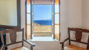 Ein Zimmer mit Meerblick durch ein offenes Fenster, ein hölzerner Stuhl und ein gerahmtes Bild an der Wand.