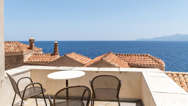 Eine Terrasse mit Tisch und Stühlen mit Blick aufs Meer, im Hintergrund Gebäude und ein klarer Himmel.