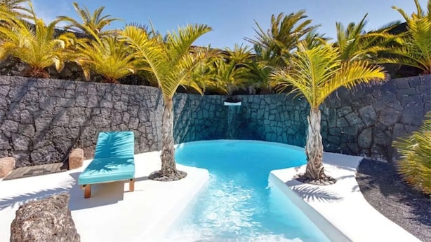 A swimming pool with a stone wall, palm trees, and a blue lounge chair. - Vacation Home 'Palacio Taro Chimida' with Private Pool (Teguise)