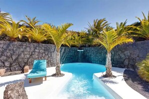 A swimming pool with a stone wall, palm trees, and a blue lounge chair.