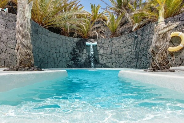 Une piscine à l’eau d’un bleu cristallin, entourée de palmiers et d’un mur de pierre.