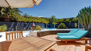 An outdoor patio with a wooden table and two blue lounge chairs, a pool, and various tropical plants. - Vacation Home 'Palacio Taro Chimida' with Private Pool (Teguise)