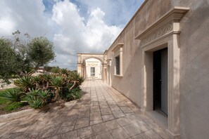 Un chemin pavé mène à un bâtiment doté d’une entrée en arc de cercle, entouré de verdure et sous un ciel dégagé.