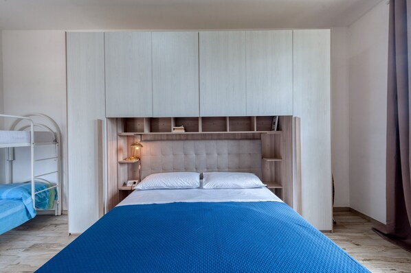 Une chambre avec un lit superposé, un grand lit recouvert d’une couverture bleue, une tête de lit en bois et des étagères intégrées.
