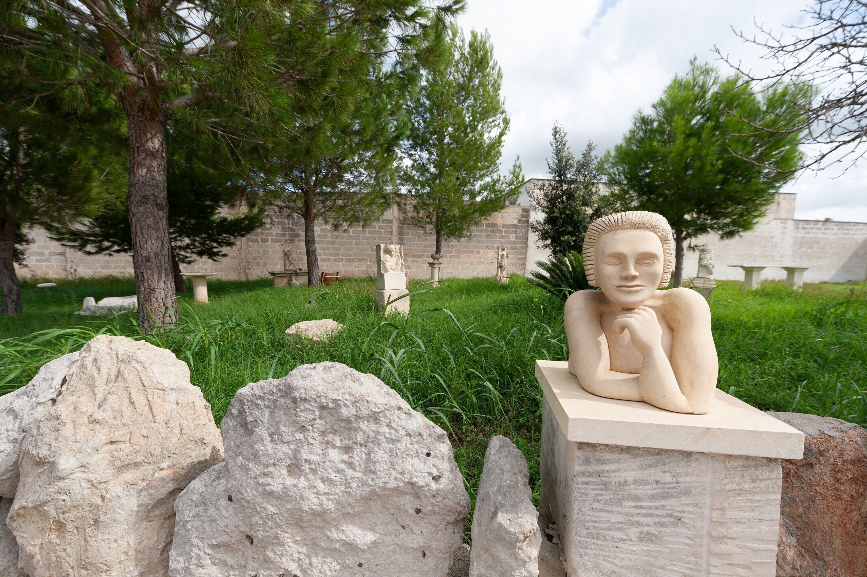 A sculpture of a reclining figure on a pedestal, surrounded by rocks and grass, with trees and a building in the background.