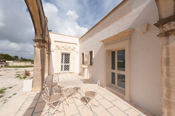 Un patio con suelo de baldosas, una mesa con sillas y un edificio con un ventanal y molduras decorativas.