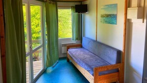 A cabin with a wooden ceiling, a single bed with a blue mattress, a TV mounted on the wall, and a window with green curtains. - Village de Gîtes Stereden - Chalet 3 - with Private Terrace and Shared Garden (Pleumeur-Bodou)